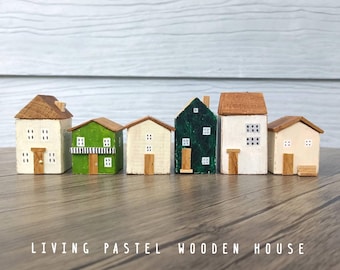 Puede incluir: Un conjunto de seis casas de madera en miniatura de varios colores, incluyendo blanco, verde y verde oscuro, cada una con un techo marrón, pequeñas ventanas pintadas y una puerta. El texto LIVING PASTEL WOODEN HOUSE aparece en la parte inferior.