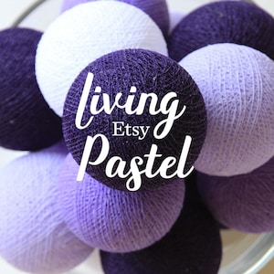 Puede incluir: Una colección de luces de bolas de algodón en varios tonos de morado, desde lavanda claro hasta violeta oscuro. Las luces están dispuestas en un cuenco y el texto "living Pastel Etsy" está superpuesto en la imagen.