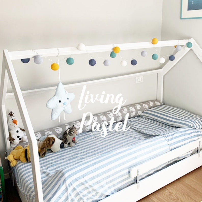 May include: White wooden bed frame with a house-shaped canopy. The bed has a blue and white striped bedding and a white star-shaped stuffed toy hanging from the canopy. The canopy is decorated with a string of white, gray, blue, and yellow cotton ball lights. The text "living Pastel" is written on the bed.