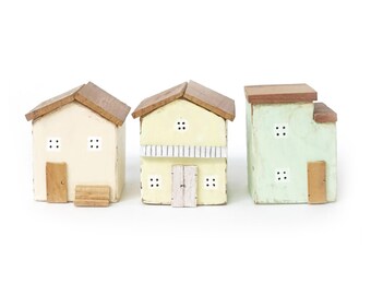 Puede incluir: Tres pequeñas figuras de casas de madera con techos planos y fachadas pintadas. La primera es de color crema con una puerta marrón, la segunda es de color amarillo pálido con una puerta central y balcón, y la tercera es verde claro con puerta marrón.