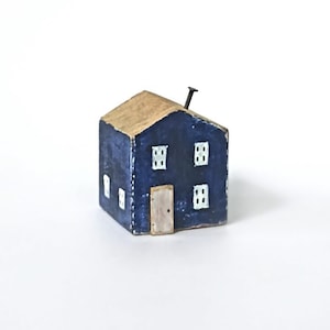 Könnte beinhalten: Eine kleine, handbemalte Holzhausfigur mit dunkelblauer Fassade, hellbraunem Dach und einem einzelnen Metallnagel, der oben herausragt. Die Vorderseite zeigt eine hellbraune rechteckige Tür und vier weiß bemalte Fenster.