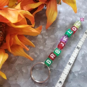 May include: A colorful beaded keychain with the letters "BRBJKLOT" spelled out. The keychain is about 3 inches long and has a silver key ring.