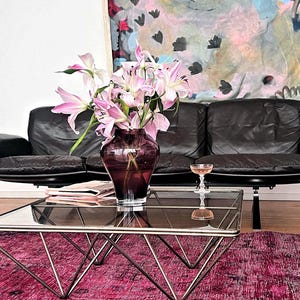 May include: A living room scene featuring a black leather sofa, a glass-topped coffee table with a geometric metal frame, and a vibrant pink rug. A vase of pink lilies sits on the table, with a glass of rose wine. An abstract painting hangs on the wall.