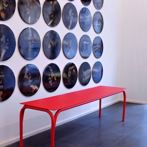 Può includere: Una vivace panca rossa con gambe curve è posizionata di fronte a un muro bianco adornato da numerose opere d'arte ovali. Le opere presentano disegni astratti in tonalità di blu, nero e marrone. Il design minimalista della panca completa l'esposizione artistica.