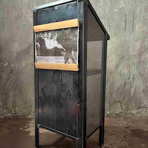 Könnte beinhalten: Ein schwarzer Metall-Präsentationsschrank mit schräger Oberseite und Gitterseitenwand. Auf der Vorderseite ist ein Schwarz-Weiß-Foto mit Holzleisten befestigt. Der Text auf dem Foto ist teilweise sichtbar.