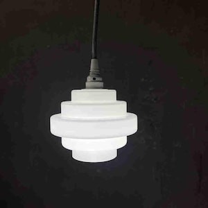 Puede incluir: Una lámpara colgante blanca de varios niveles con un cable negro. La lámpara tiene un diseño escalonado, con cada nivel ligeramente más pequeño que el anterior. Cuelga sobre un fondo oscuro.