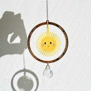 May include: A decorative suncatcher featuring a smiling yellow sun within a gold-colored ring. The ring has text in French and the years 2024-2025. A clear crystal ball hangs below, suspended by a chain. The suncatcher is hanging against a white background.
