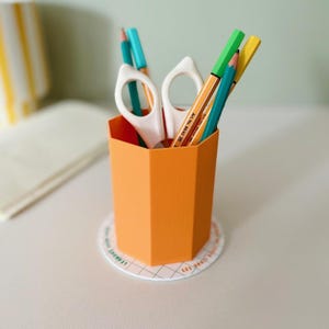 May include: An orange, geometric pencil holder containing pencils and white scissors. The pencils are various colours, including green, yellow, and teal. The holder sits on a round coaster with text.