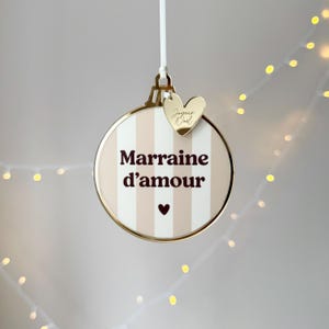 Op de afbeelding: Een ronde kerstversiering met een goudkleurige rand en een gestreepte beige en witte achtergrond. De versiering heeft de woorden "Marraine d'amour" in donkerbruin, een klein hartje en een goudkleurige hartvormige bedel met tekst. Een wit lint is bevestigd.
