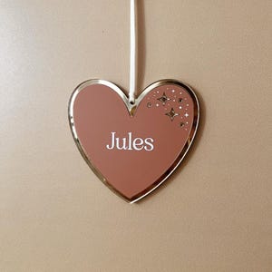 Könnte beinhalten: Ein herzförmiger Schmuckanhänger mit goldfarbenem Rand und einem staubrosafarbenen Mittelteil. Das Wort "Jules" ist weiß gedruckt. Dekorative Sterne befinden sich in der oberen rechten Ecke. Ein weißes Band ist zum Aufhängen befestigt.