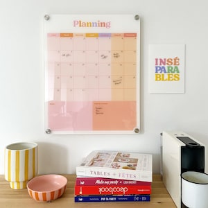 Könnte beinhalten: Ein klarer Wandkalender aus Acryl mit dem Wort "Planning" in Pastellfarben. Der Kalender hat ein rosa-orangenes Farbschema. Ein Stapel Bücher, eine gestreifte Schüssel und eine gelb-weiß gestreifte Vase befinden sich auf einer Holzoberfläche.