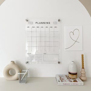 May include: A clear acrylic wall planner with the word "PLANNING" at the top. The calendar grid is divided into days and includes a to-do list and notes section. A white heart-shaped art print and decorative items are also present.