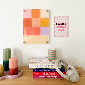 May include: A colorful wall-mounted planner with squares for each day of the week, and a pink sign that says "S'aimer comme jaja". There are also candles, books, a striped mug, and headphones on a wooden surface.
