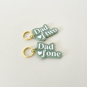 May include: Two keyrings with the text "Dad of two" and "Dad of one" in white on a sage green background. Each keyring has a gold-coloured ring. A thoughtful gift for fathers.