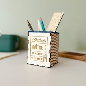 Peut inclure: Organisateur de bureau en bois avec l'inscription "Meilleur Maître du Monde Entier" gravée. Il contient des stylos, des crayons et une petite règle. Les côtés présentent un motif rayé.