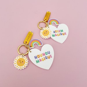 May include: Two keychains with gold-colored hardware, featuring a white heart-shaped charm with the text "ATSEM MAGIQUE" and "NOUNOU MAGIQUE" in colorful letters. Each keychain has a rainbow, a smiling sun, and a rectangular tag with a name.