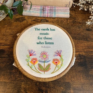 May include: A white embroidery hoop with a wooden frame, featuring a quote by Shakespeare: "The earth has music for those who listen." The quote is embroidered in green thread, and the hoop is decorated with colourful embroidered flowers.