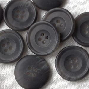 Puede incluir: Un conjunto de botones grises oscuros con cuatro agujeros cada uno, dispersos sobre una superficie de tela blanca. Los botones tienen un sutil patrón de vetas de madera. Adecuados para coser o manualidades.