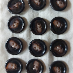 May include: A collection of twelve dark brown buttons with a marbled, light brown center. Each button has four holes for sewing. The buttons are arranged on a white fabric with the word "voy" faintly visible.