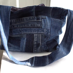 May include: A denim tote bag constructed from various shades of blue jean fabric. The bag features a patchwork design with a long strap for shoulder or hand carry. The bag is sitting on a white surface.