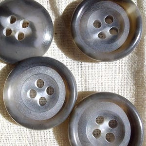 May include: Four circular buttons with a smooth, semi-translucent gray finish. Each button features a recessed center with four evenly spaced holes for attachment. The buttons have a polished surface that reflects light, creating subtle highlights.