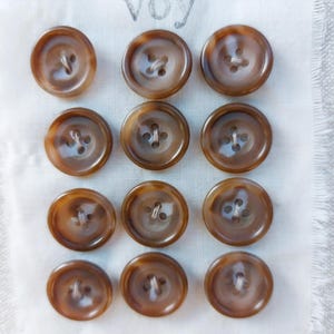 May include: A collection of twelve brown, circular buttons with four holes, displayed on white fabric. The buttons have a marbled effect. The word "voy" is printed in grey above the buttons.