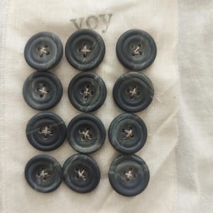 May include: A collection of twelve dark gray buttons with a marbled effect, each featuring four holes and beige thread. The buttons are arranged on a white fabric, with the word "voy" faintly visible in the upper left corner.