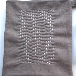 May include: A square, grey-brown fabric item featuring a rectangular area of white, hand-stitched detail. The stitching forms a wavy pattern. A small loop extends from the top edge. The item is likely a kitchen accessory, such as a pot holder or oven mitt.