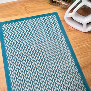 Puede incluir: Un esténcil rectangular con un borde azul y un patrón blanco repetitivo en forma de hoja. El esténcil se coloca sobre una superficie de madera de color marrón claro. Otros objetos en el fondo incluyen una bola de musgo decorativa y un objeto de plástico blanco.