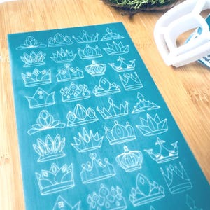 May include: A teal blue sheet with white outlines of various crown designs. The sheet is on a wooden surface.
