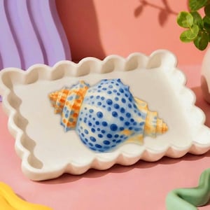 Peut inclure: Un plateau rectangulaire en céramique blanc cassé avec un bord festonné présentant une illustration centrale de coquillage. Le coquillage est peint avec un dégradé d'orange et de jaune aux extrémités et un motif de points bleus au centre.