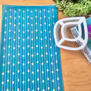 May include: A blue stencil with a repeating pattern of white lines and circles. The stencil is on a wooden surface next to a white plastic stencil.