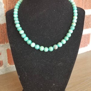 May include: A turquoise beaded necklace on a black velvet display stand. The beads are round and have a slightly irregular shape.