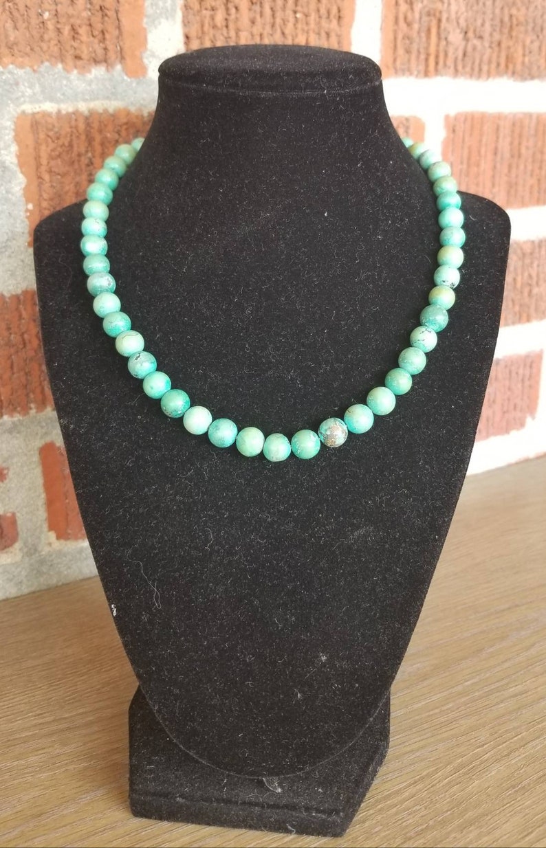 May include: A turquoise beaded necklace on a black velvet display stand. The beads are round and have a slightly irregular shape.