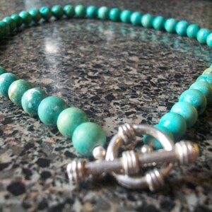 May include: A turquoise gemstone necklace with a silver clasp. The necklace is made of round beads and is approximately 18 inches long.
