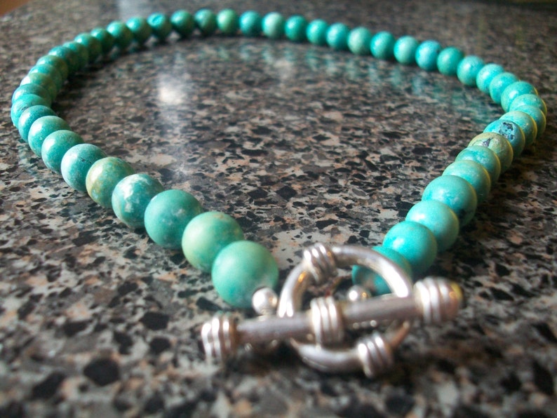 May include: A turquoise gemstone necklace with a silver clasp. The necklace is made of round beads and is approximately 18 inches long.