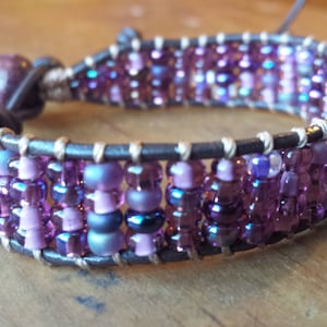 May include: A purple and grey beaded bracelet with a brown leather wrap. The bracelet is made with small, round beads and has a simple design.