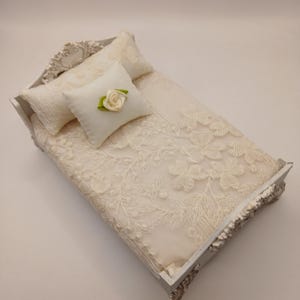 May include: A miniature bed frame in a light gray finish with ornate carved detailing on the headboard and footboard. The bed is dressed in cream-colored bedding featuring an intricate floral lace overlay, two rectangular pillows, and a small square accent pillow adorned with a single cream fabric rose with green leaves.