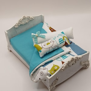 May include: A miniature white bed with a turquoise blanket and pillows. The pillows and blanket feature a cat pattern in green, blue, and brown. The bed frame has ornate detailing.