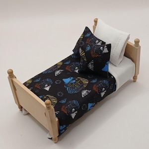 May include: A miniature wooden bed with a black patterned duvet cover and pillow set. The duvet cover and pillow feature a repeating pattern of colourful fish. The bed also includes a white pillow and a white sheet.