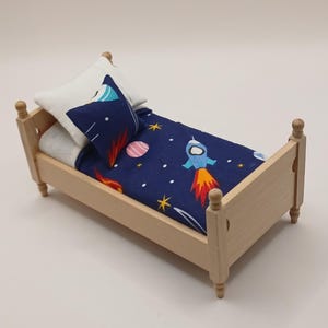 May include: A miniature wooden bed with a navy blue duvet cover and pillow set. The duvet cover features a rocket ship, planets, and stars design. The pillow is white with a blue and grey design. The bed frame is light brown.