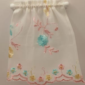 May include: A sheer white curtain panel with embroidered floral designs in pastel colors. The embroidery features pink, yellow, and blue flowers and vines. The curtain is gathered at the top and held by two white ball-shaped finials.