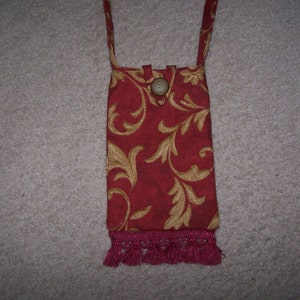 Könnte beinhalten: Eine rote und goldene Stofftasche mit Knopfverschluss und einem rosa Fransenbesatz. Die Tasche hängt an einem Riemen.
