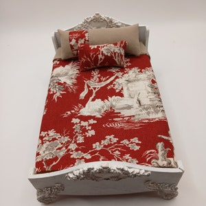 May include: A miniature white bed with ornate detailing. The bed features a red and white toile patterned comforter and matching decorative pillows. Two beige pillows and a small red pillow complete the bedding set. The bed frame has decorative carvings.