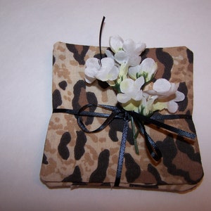 May include: A small gift box wrapped in brown fabric with a leopard print pattern. The box is tied with a black ribbon and decorated with white flowers.