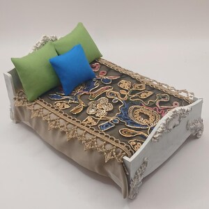 May include: A miniature bed with a white frame and ornate detailing. The bed is adorned with a patterned dark gray blanket, a tan bed skirt, and three pillows in green and blue. The bed frame has decorative carvings.