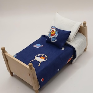 May include: A miniature wooden bed with a navy blue comforter featuring a space-themed pattern of rockets, planets, and UFOs. It includes a white pillow and a matching pillow with a rocket design. The bed frame is light-colored wood.