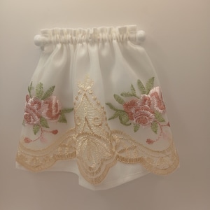 May include: A white, sheer curtain valance with floral embroidery. The valance features pink and green embroidered flowers and a scalloped, beige lace trim. The top has a gathered design with small white ball accents.