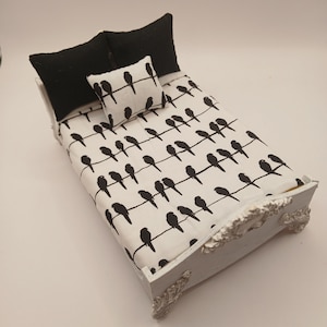 May include: A miniature white bed with a black and white bird print on the bedding and a small pillow. Two black pillows are at the head of the bed. The bed frame has ornate detailing.