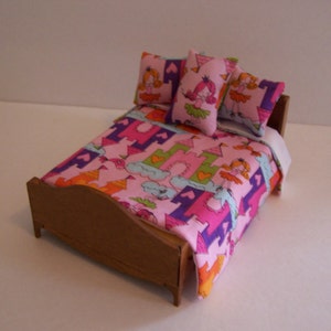Può includere: Un letto in legno in miniatura con un set di biancheria da letto a tema castello rosa e viola. La biancheria da letto presenta un motivo ripetitivo di castelli, nuvole e cuori. Ci sono tre cuscini con un design di principessa e castello.